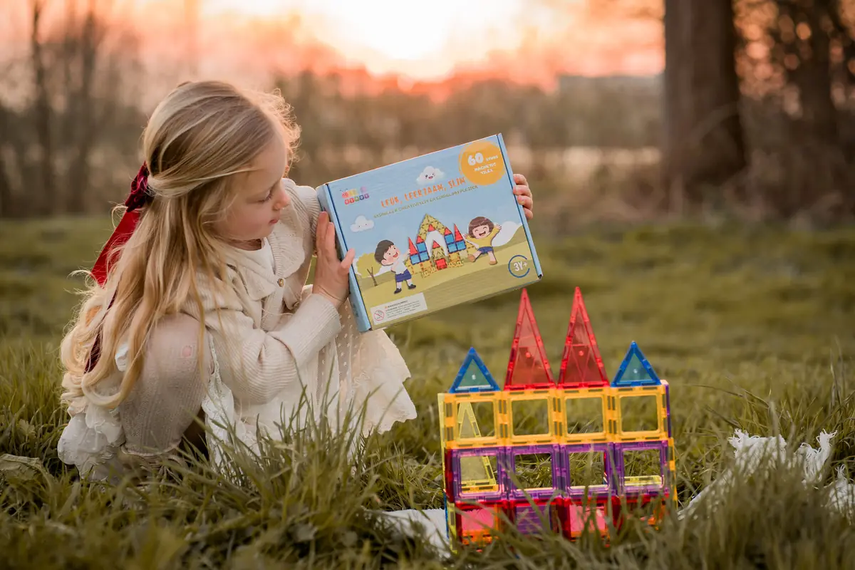 Meisje zit in het gras naast een bouwwerk en een doos magnetische tegels